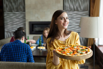 Young beautiful girl poses with a pizza in a cafe against the background of her friends.