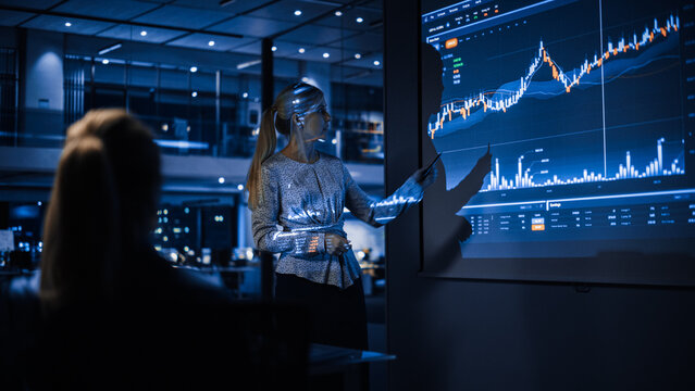 Business Conference Meeting Presentation: Businesswoman Does Financial Analysis Talks To Group Of Businessspeople. Projector Screen Shows Stock Market Data, Investment Strategy, Revenue Growth