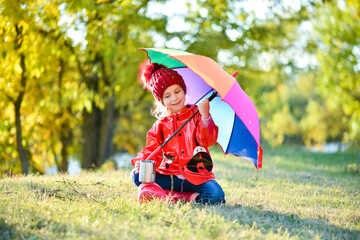 A smiling girl in a red hat and a red jacket walks with a rainbow umbrella in nature. Yellow leaves in the background. Autumn. Atmosphere