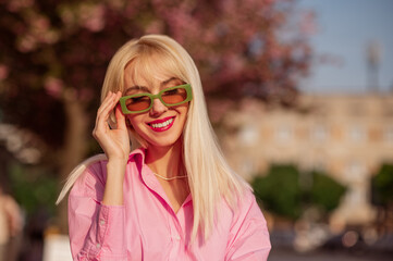 Outdoor fashion portrait of happy smiling blonde woman wearing trendy green rectangle sunglasses,...
