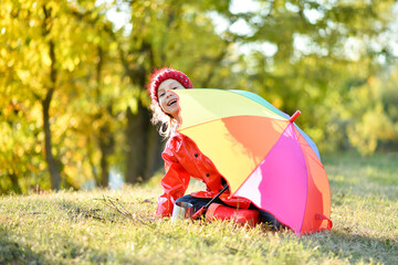 A smiling girl in a red hat and a red jacket walks with a rainbow umbrella in nature. Yellow leaves...