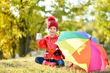 A smiling girl in a red hat and a red jacket walks with a rainbow umbrella in nature. Yellow leaves...