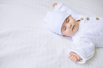 a newborn baby sleeps in a crib in a white warm jumpsuit and a hat on a white insulated cotton bed,...