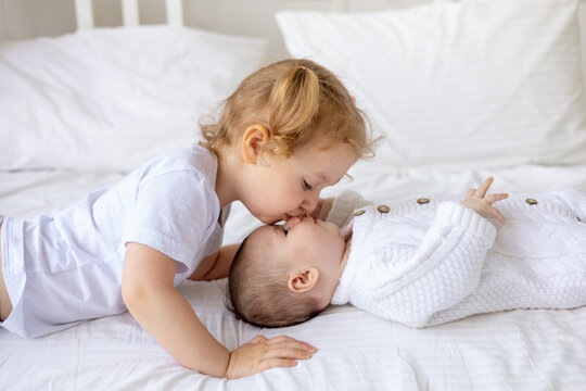 sister gently hugs and kisses a newborn baby in a crib on a white insulated cotton bed, two children in the family brother and sister