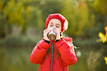 Teenager girl in a red hat and a red jacket drinking a cup mug enjoys nature. Yellow leaves in the...