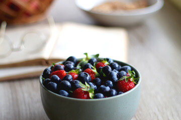 Bowl of strawberries and blueberries, open books with reading glasses, plate of chocolate chip cookies and basket of apples one the table. Selective focus.