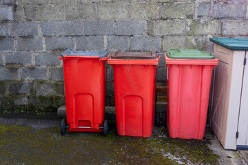 Obraz premium Red small wheelie recycling plastic bins in a back yard of a house by a wall. Waste disposal industry.