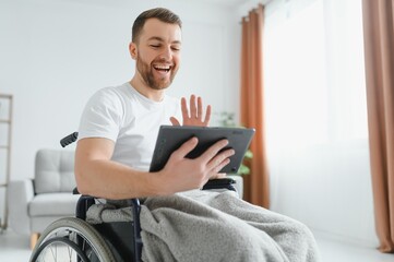 Man in wheelchair using tablet computer at home. Positive retired male with physical disability browsing web on touch pad, watching video online indoors.