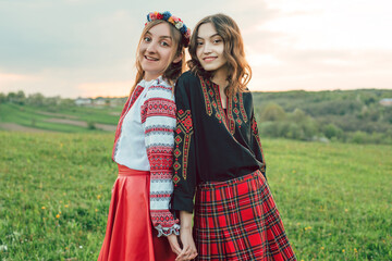two girls in traditional ukrainian clothes - vysyvanka holding hands and smiling at camera in the field during sunset. 