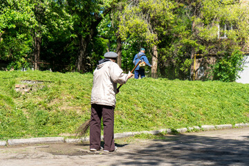 Old woman sweeping the street in the city. Man in the background mows the grass