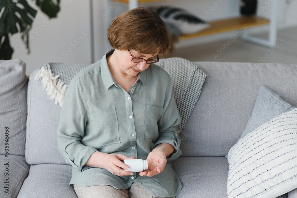 Sticker Eldery woman sitting on the sofa and taking pills at home. Pensioner with medicaments