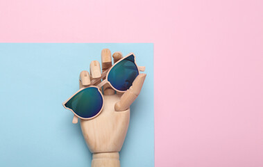 Wooden hand holding stylish sunglasses on blue pink pastel background