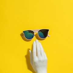 Minimal summer layout. Plastic hand reaching for stylish sunglasses on a bright yellow background....