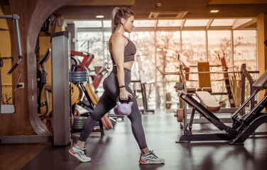 Fototapeta na wymiar Fit woman practices lunges with kettlebells in her hands in the gym. Leg workout