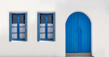 Blue door and two windows with open shutters on white wall. Greek island house front view