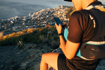 Close up woman hiker in sportsclothes checking smartphone while sitting on the top of the mountain...