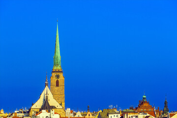 Fototapeta premium Gothic temple tower with a clock, roofs of houses with Christian crosses and modern antennas under an ultramarine sky in the morning sunlight in the city of Pilsen in the Czech Republic
