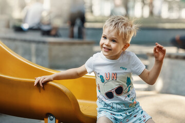 A cheerful, smiling, likeable little boy on the playground on a yellow slider. The child's hair is...