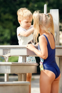 A happy boy and a cute girl in a blue bathing suit play with a water tap in a city park. Vertical