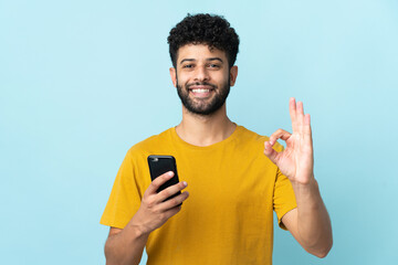 Young Moroccan man isolated on blue background using mobile phone and doing OK sign