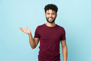 Young Moroccan man isolated on blue background holding copyspace imaginary on the palm to insert an...