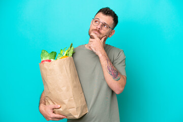 Young Brazilian man holding a grocery shopping bag isolated on blue background having doubts