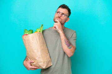 Young Brazilian man holding a grocery shopping bag isolated on blue background and looking up