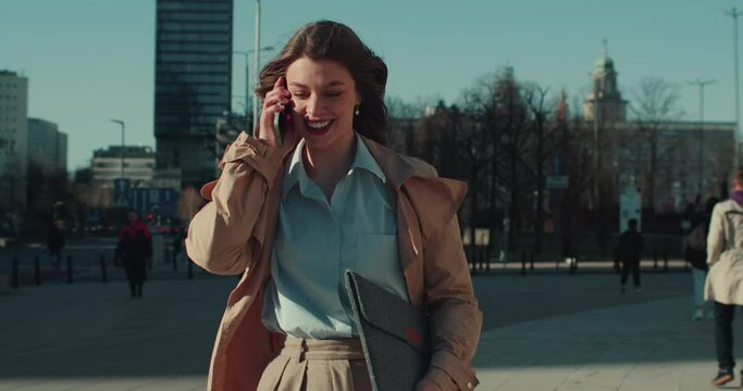 Happy young beautiful business woman picks up phone call, talks walking along windy sunny city square slow motion.