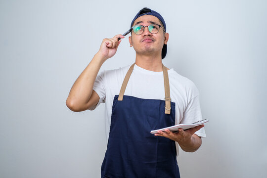 Asian barista man in blue apron taking order, writing on menu book list, thinking with pen on head - Powered by Adobe