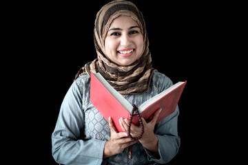 Portrait of smiling beautiful Muslim young woman wearing hijab scarf, reading Muslim book Quran (holy Koran book of Islam) in her hand on dark black background.