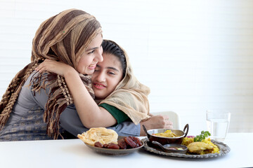 Adorable Pakistani Muslim girl sits at kitchen table with traditional Islamic halal food, kid with hijab hugs mother, warm love in family of mom and daughter, happy Islamic family on white background. - obrazy, fototapety, plakaty