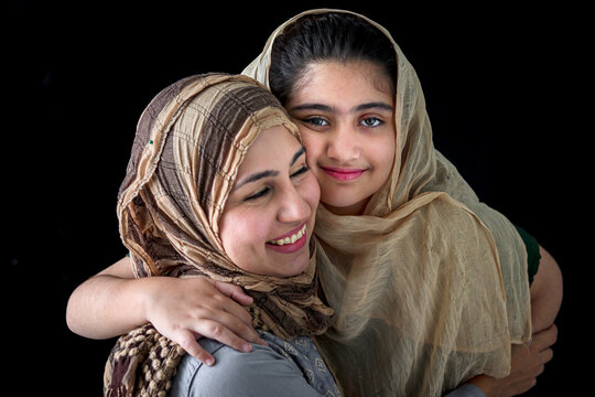 Adorable smiling Pakistani Muslim girl with beautiful eyes wearing hijab, hugging mother, warm love in family of mom and daughter, happy lovely Islamic family on dark black background.