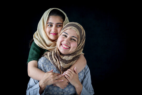 Portrait of smiling Pakistani Muslim woman with daughter, adorable beautiful eyes kid girl with hijab hugs mother, warm love of mom and daughter, happy lovely Islamic family on dark black background.