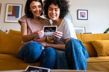 Smiling multiracial lesbian pregnant couple looking baby ultrasounds at home sitting on the couch.