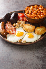 Full English breakfast with bacon, sausage, fried egg, baked beans, hash browns and mushrooms closeup in the plate on the table. Vertical