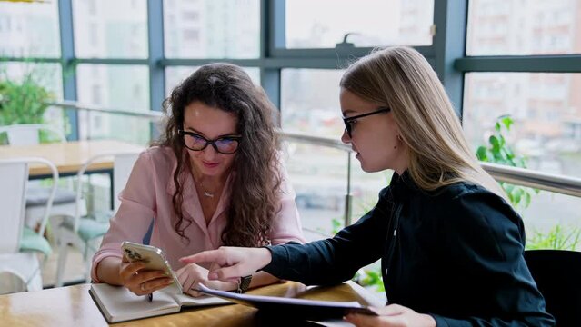 Two young women look at smartphone and discuss something smiling. Female office workers talk about ideas.