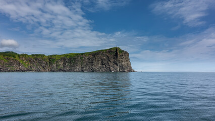 The rocky coast of Kamchatka against the background of blue sky and clouds. Steep slopes, green vegetation on the hills. Ripples on the surface of the Pacific Ocean. Avacha Bay