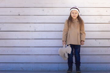 Caucasian little girl of 5 6 years with teddy bear in hand looking at camera on white colored wooden background in coat and hat