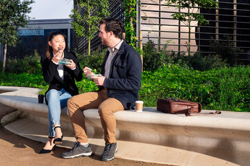 asian woman talking and eating a salad with her caucasian partner sitting in a park next to the...