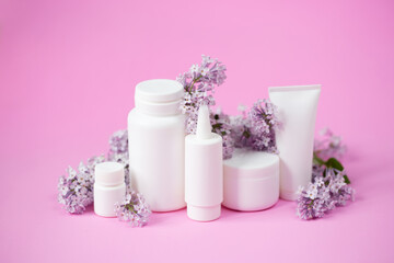 Face and body skin care. A set of cosmetic creams and balms in white tubes and cans on a coral background with sprigs of lilac flowers. Spa treatments for home care. Home rejuvenation and moisturizing