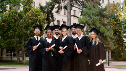 Group of multiethnic graduates students in the college park posing excited with large smile in front of the camera with diplomas in hands they wearing graduation caps