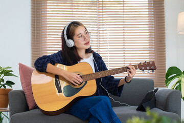 Happy Asian glasses girl playing acoustic guitar and listening music from tablet computer in living...