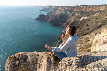 Woman tourist enjoying the sunset over the sea mountain landscape. Sits outdoors on a rock above the sea. She is wearing jeans and a blue hoodie. Healthy lifestyle, harmony and meditation