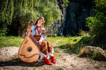Ukrainian musician in authentic national dress sits by a rock. Ukrainian woman with a bandura...