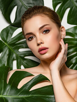 Portrait of a beautiful young girl with clean and healthy skin. Clean face of a white young girl close-up. Beautiful woman touches her face with her hand. Model with green leaves near the body. Beauty