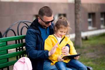 A cute girl and her dad sit on a bench with a gadget and communicate online. It's spring and warm outside. Holidays with the family. Communication online. Outdoors. Communication. Technology.