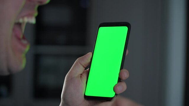 Happy millennial man watch on mobile phone with green screen at home and received amazing news on phone, online casino bet winner, showing yes gesture and screaming yeah.