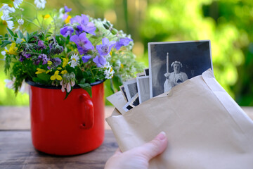 female hands holding paper envelope with old photographs, bouquet of wild flowers on table wooden...