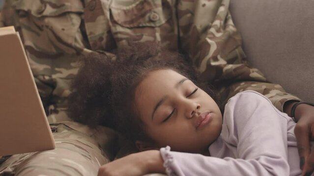 Slowmo of unrecognizable Black woman in military uniform reading to her pretty little daughter napping on her laps on sofa at home