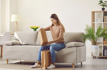 Happy customer takes box out of paper bag. Smiling young woman sitting on sofa at home and...